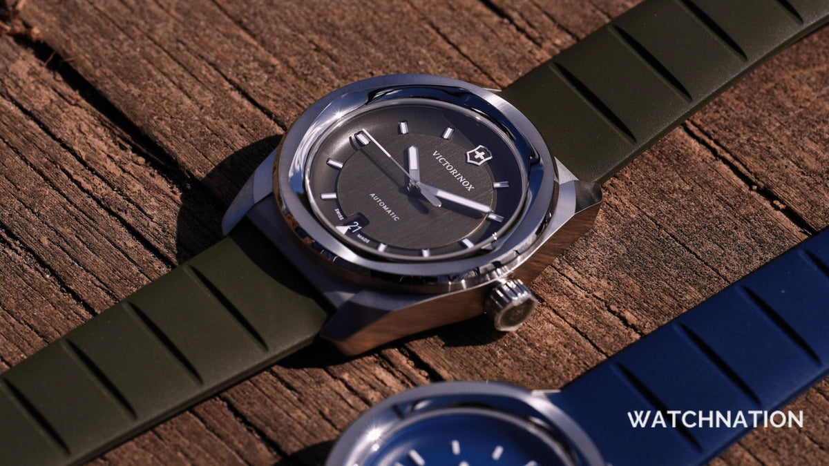 A close-up shot of a Victorinox automatic watch with a dark grey dial and an olive green rubber strap, resting on a textured wooden surface next to a blue strap version.