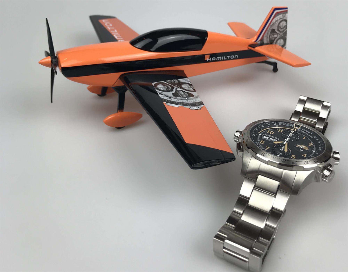 Men's steel chronograph watch with black dial and bracelet, photographed with an orange airplane model