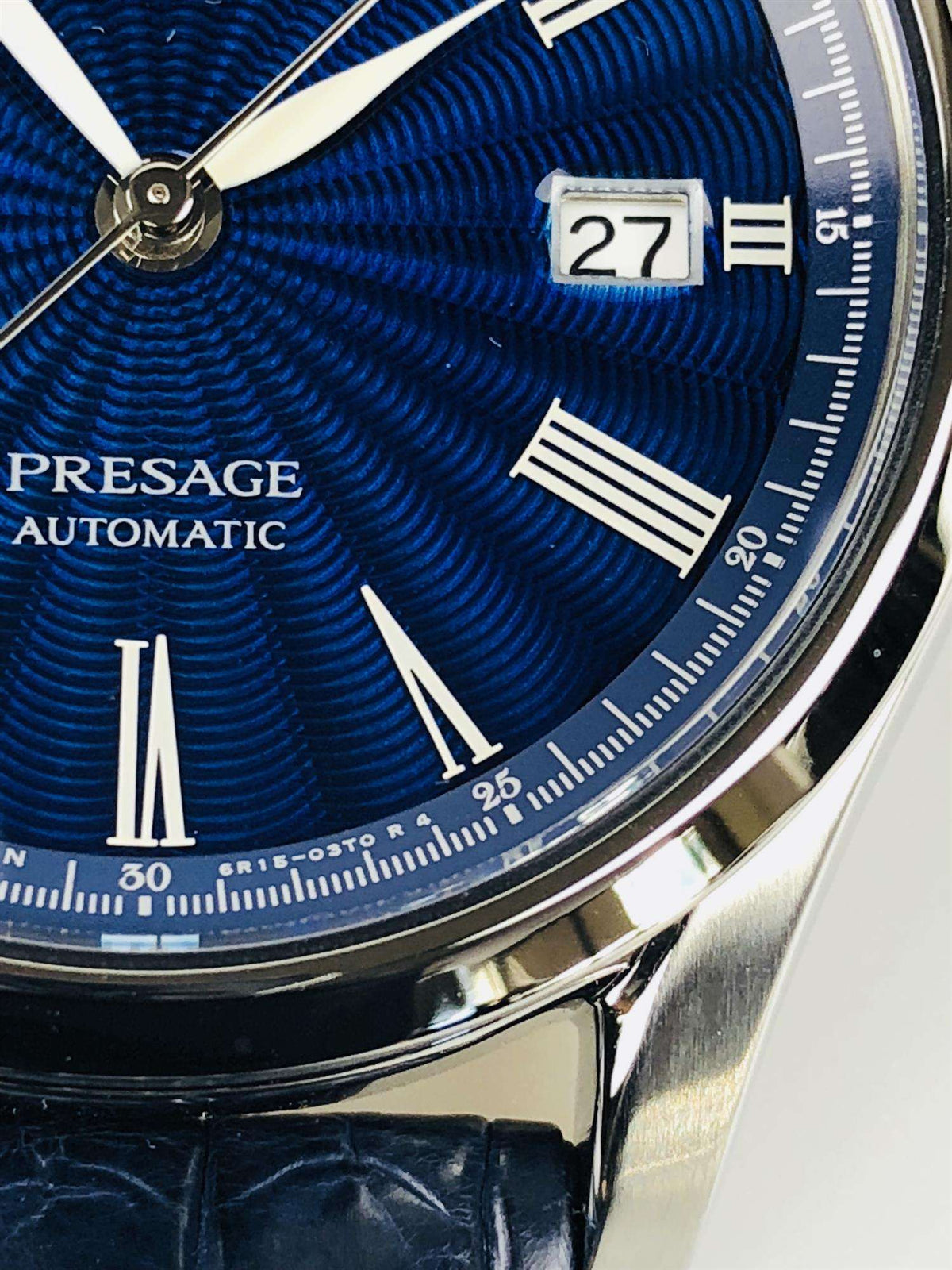Men's automatic wristwatch close-up: blue dial texture, date window at 27, silver bezel