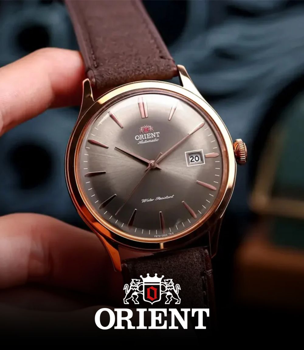 Hand holding an Orient Automatic dress watch with a rose gold-toned case, sunburst grey dial, date window, and brown leather strap, featuring the Orient logo at the bottom