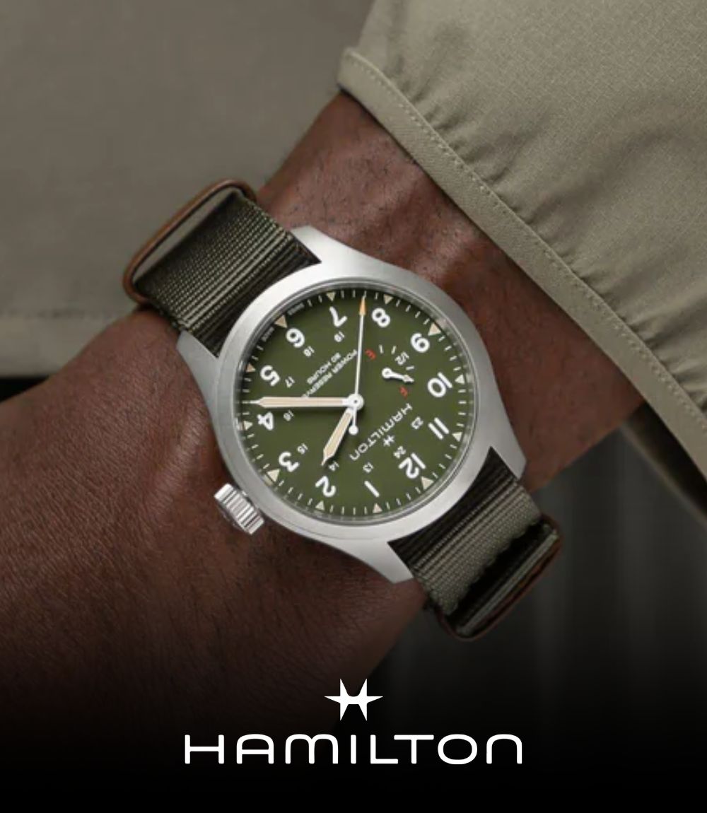 Close-up of a Hamilton field watch worn on a wrist, featuring a matte silver case, olive green dial with Arabic numerals, and an olive green NATO fabric strap, featuring the Hamilton logo at the bottom