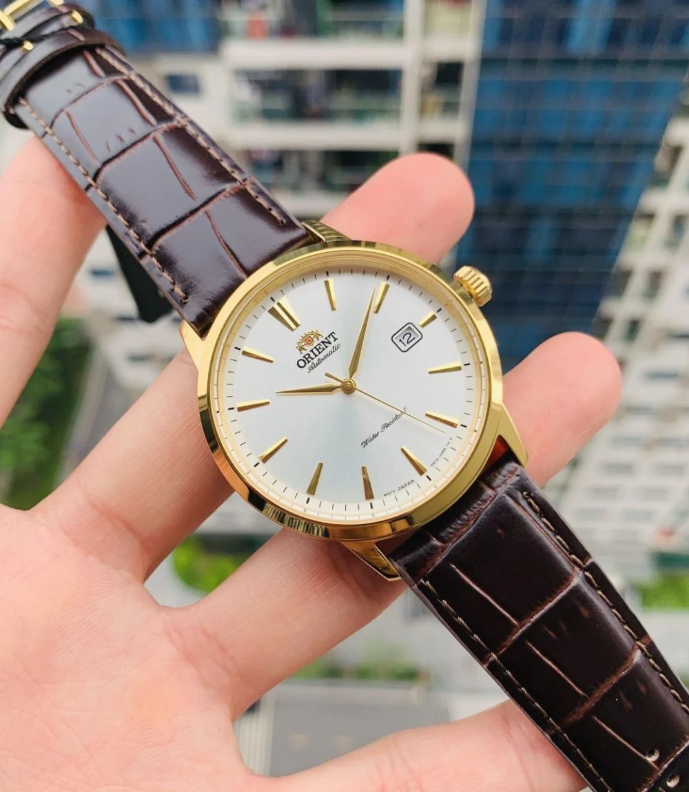 Orient Symphony gold watch featuring a white dial, gold-tone case, and brown leather strap held in hand