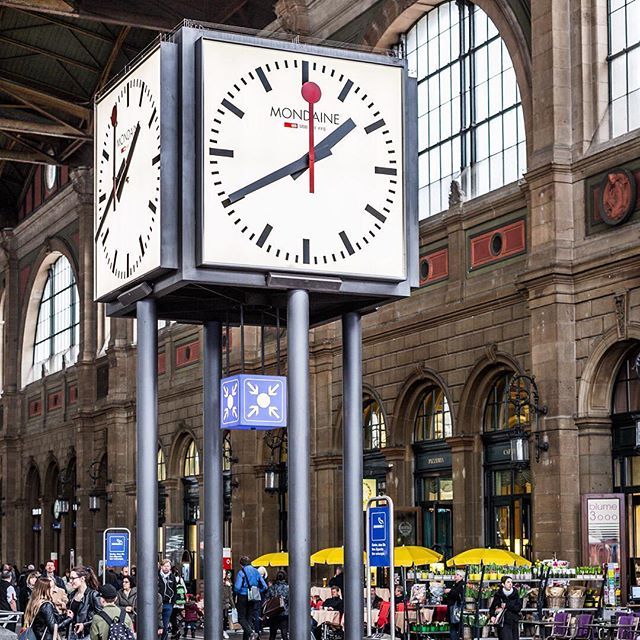 Large Mondaine Swiss railway clock mounted on pillars inside a busy train station concourse