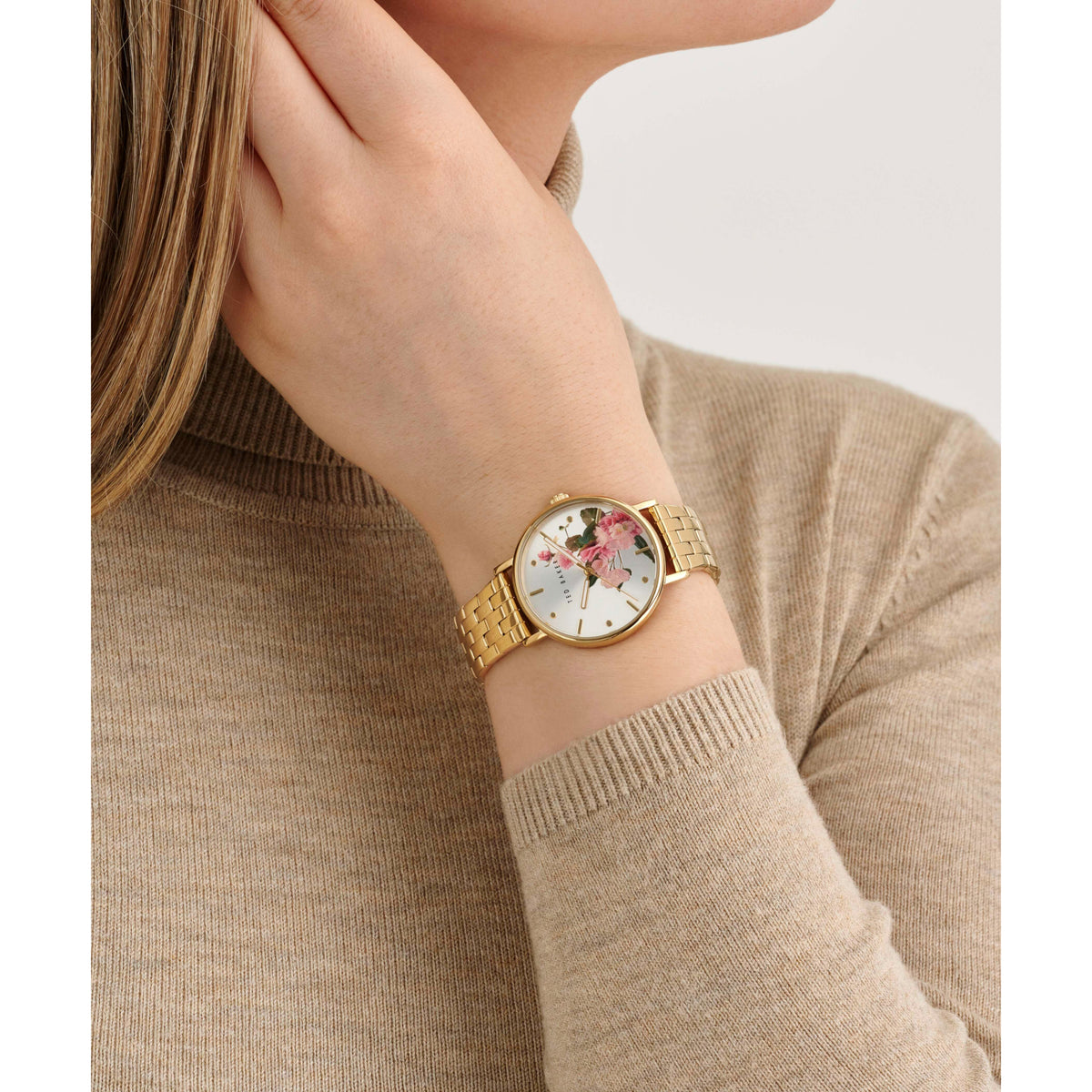 Gold-tone watch on wrist with white floral face and beige sweater sleeve