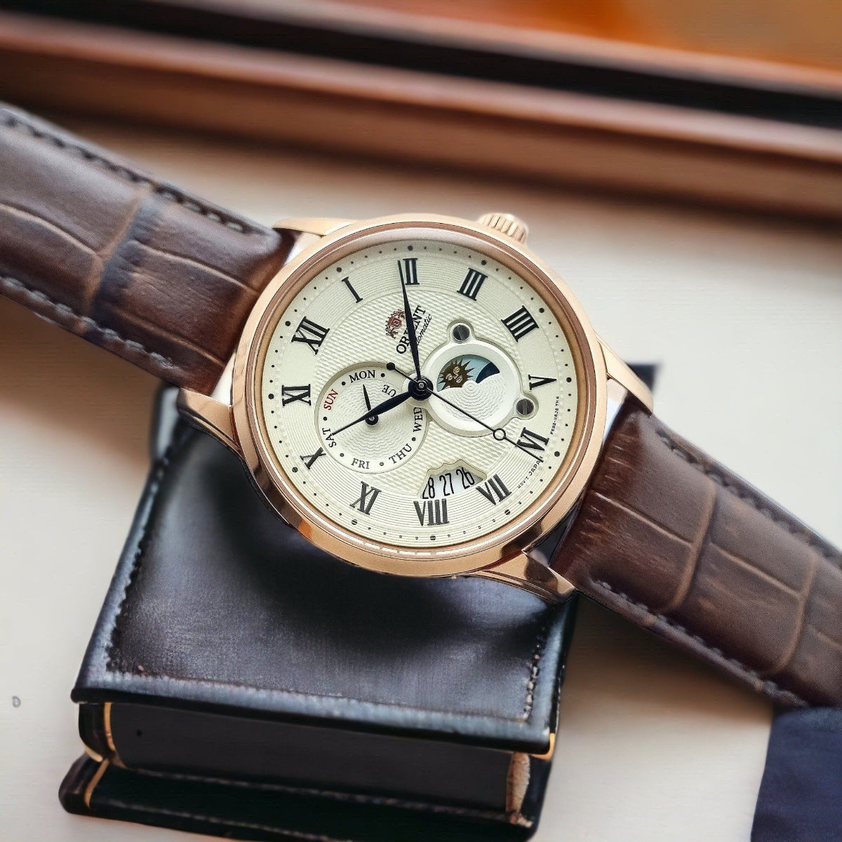 Orient Bambino Sun & Moon RA-AK0007S30B mens automatic watch featuring a champagne dial with brown leather strap resting on a black accessory box