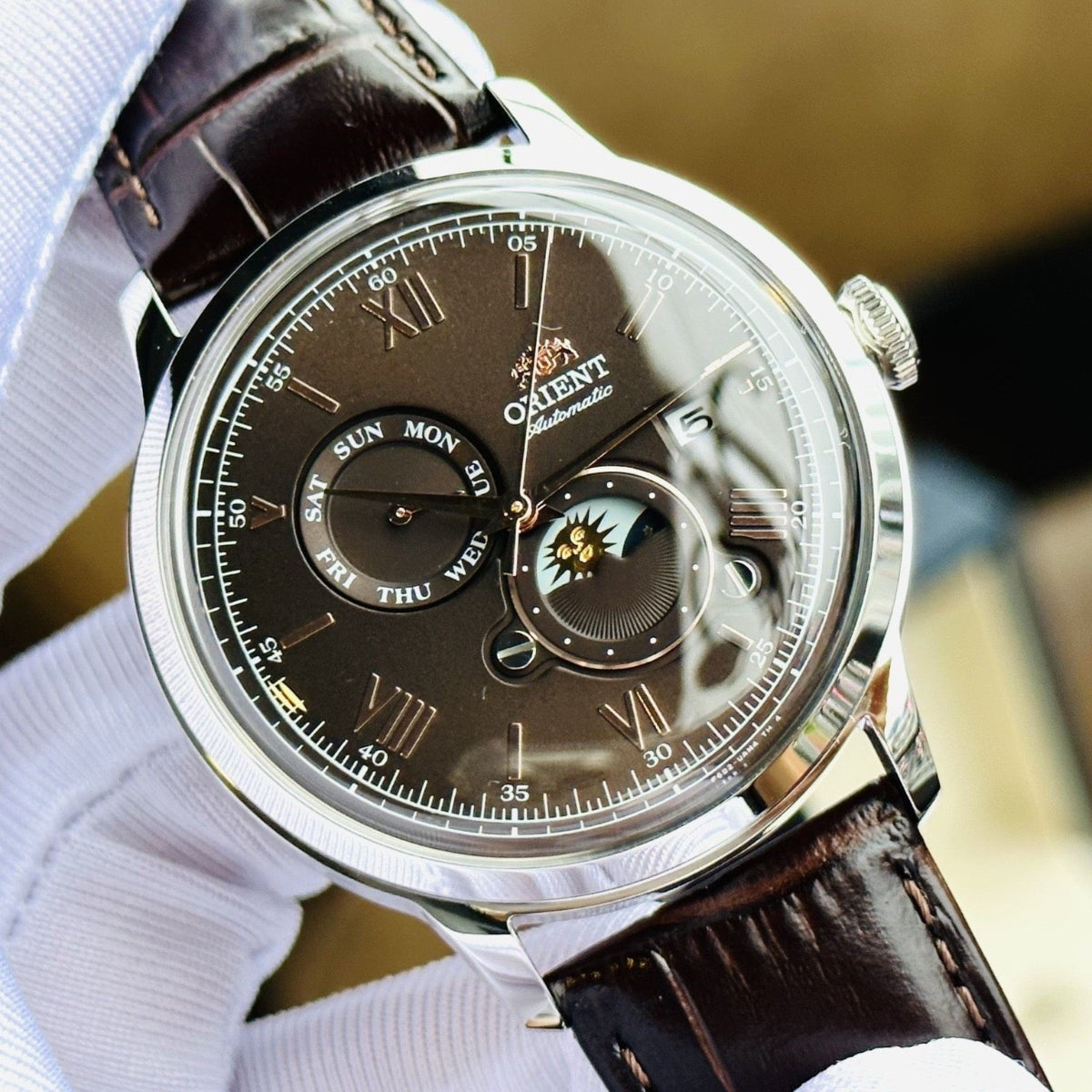 Orient Bambino Sun & Moon RA-AK0804Y30B automatic watch close-up with brown dial, sun-moon subdials, and dark brown leather strap