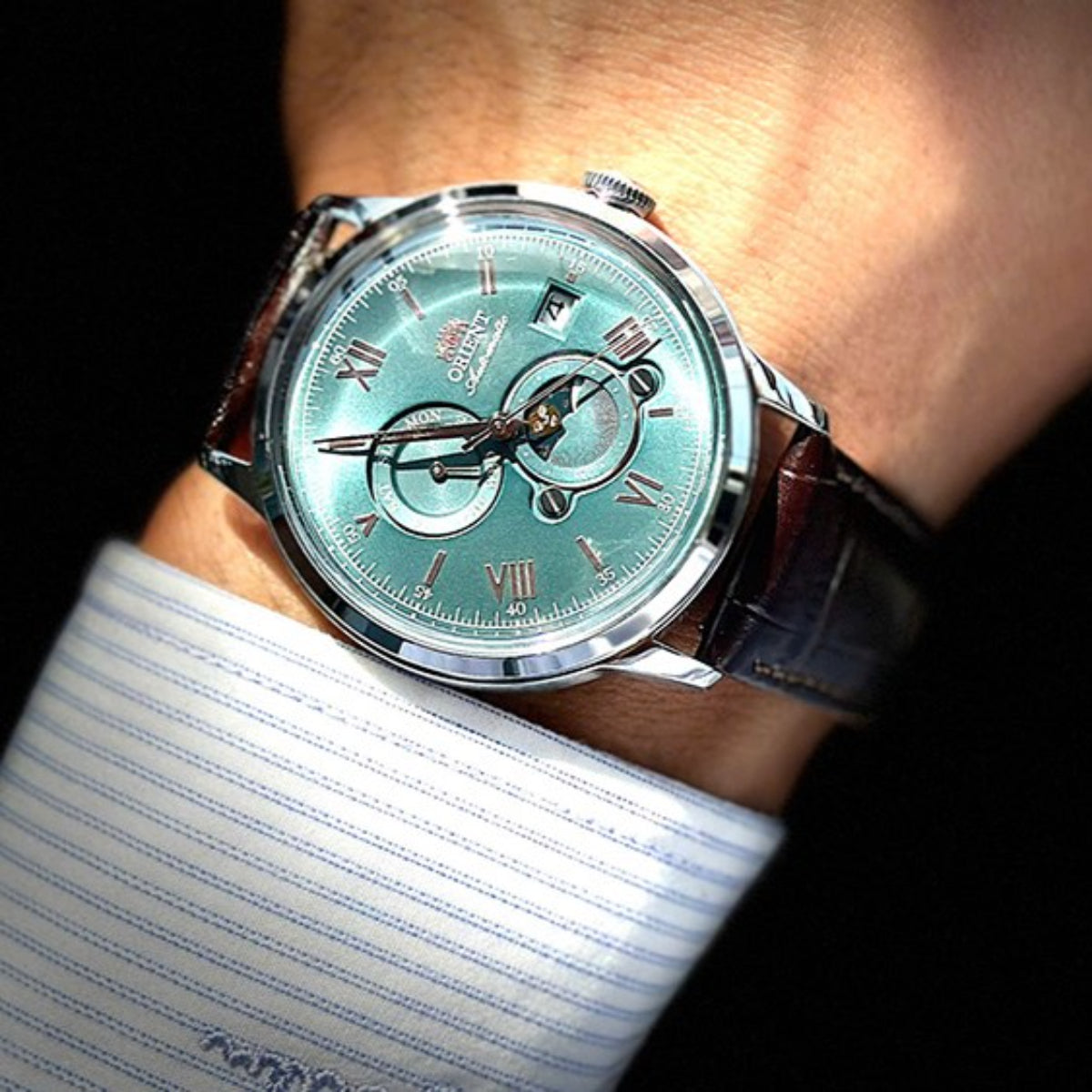Automatic watch on wrist with teal dial and brown leather strap, roman numerals and subdials