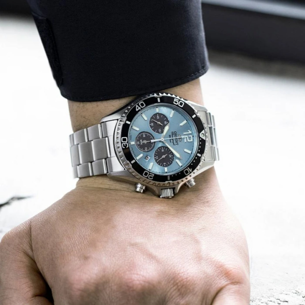 Men's wristwatch with blue chronograph dial and stainless steel bracelet on bare wrist.