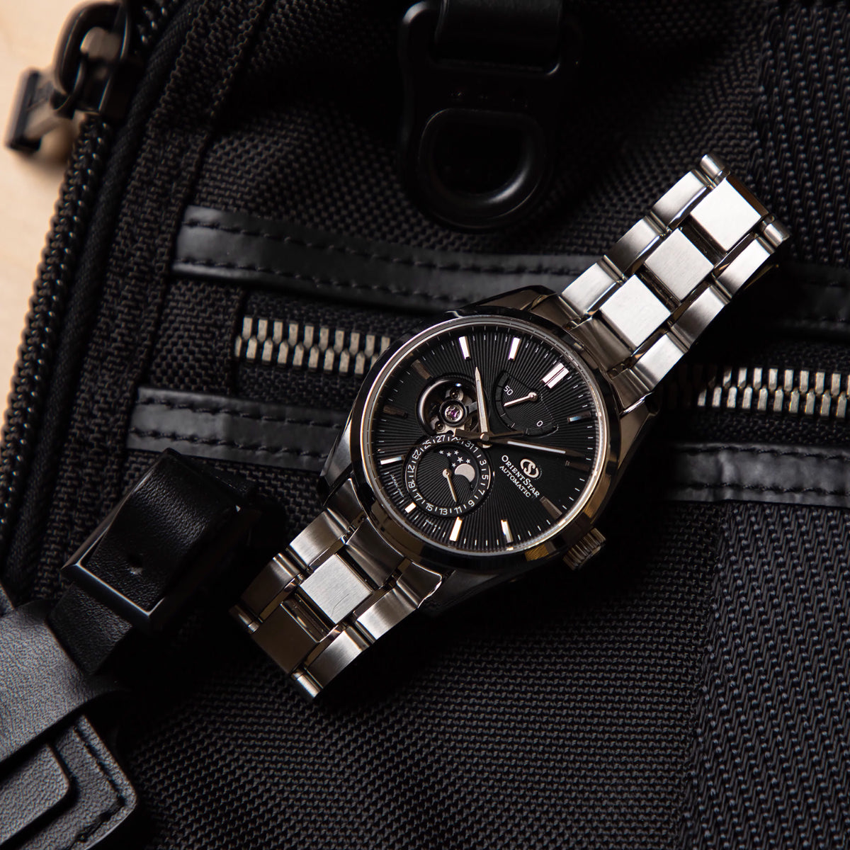 Steel moon phase watch with black dial on dark fabric; bracelet and bezel visible