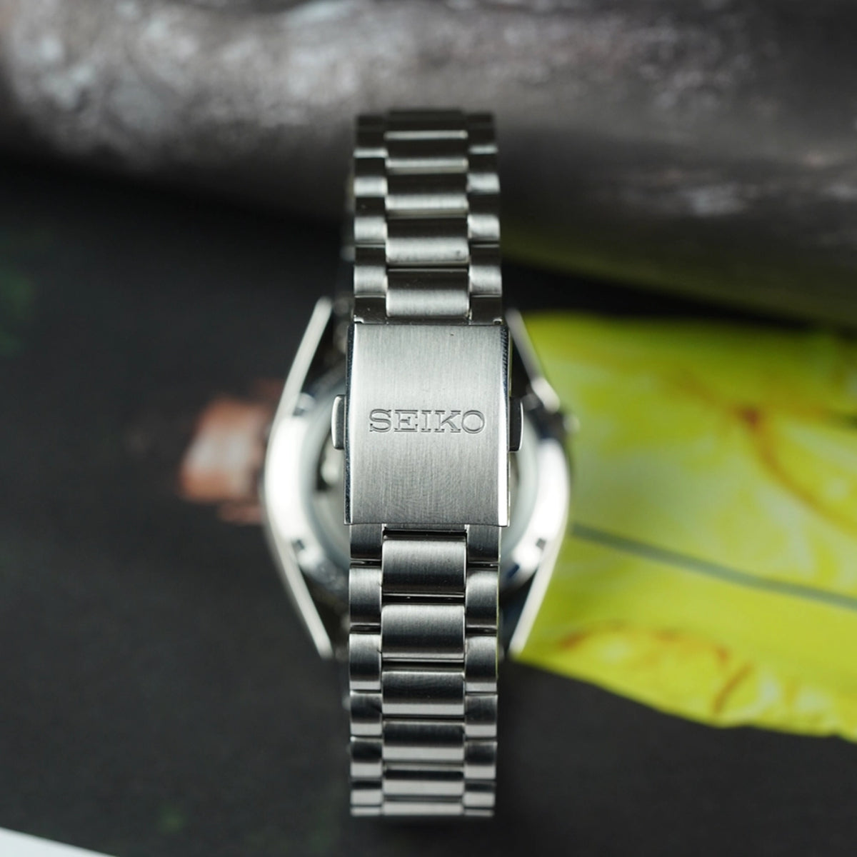 Seiko 5 SNXS bracelet lying flat, showcasing the brushed steel finish and the brand-engraved folding clasp with a blurred background