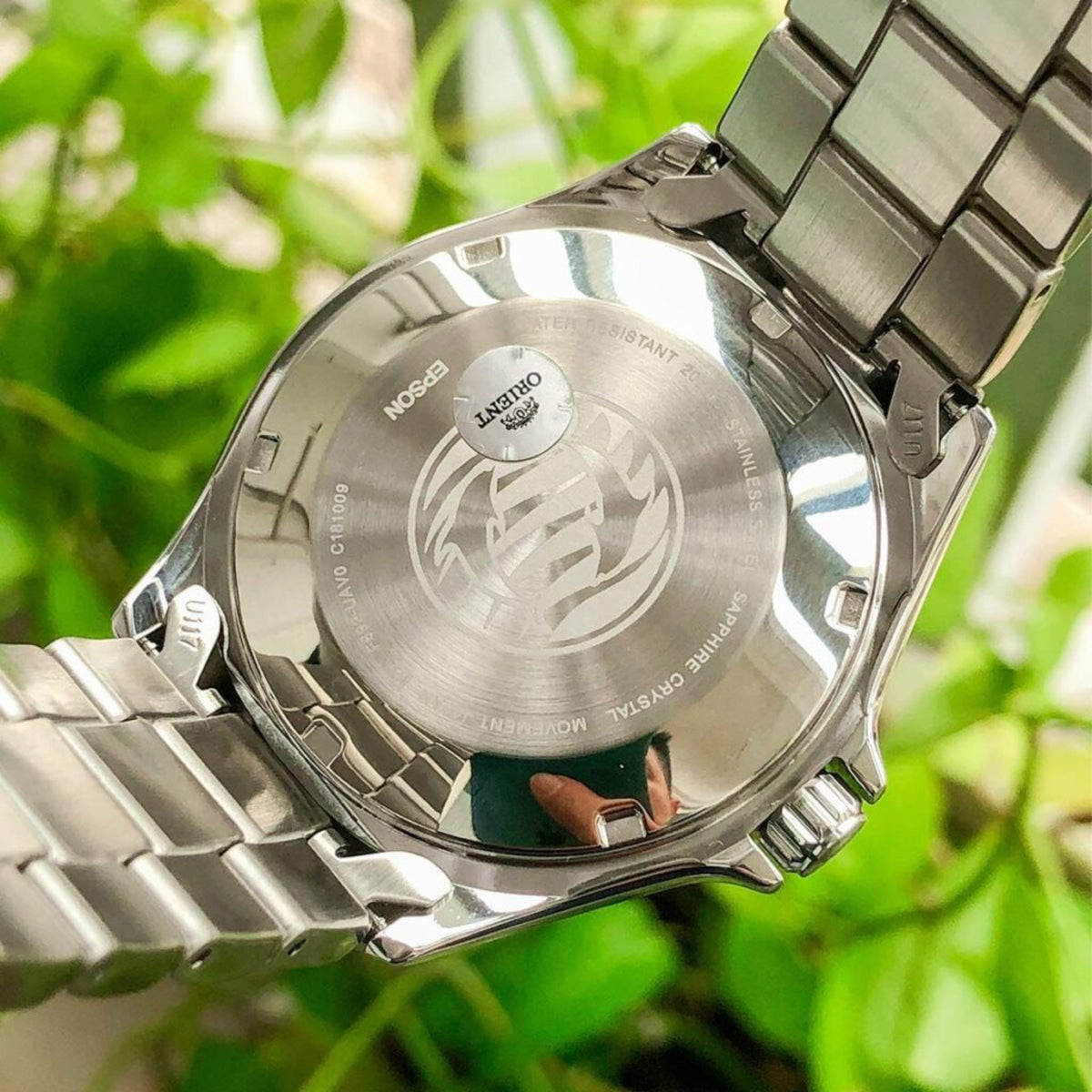 Close-up of a silver watch with brand logo against a blurred green leafy background