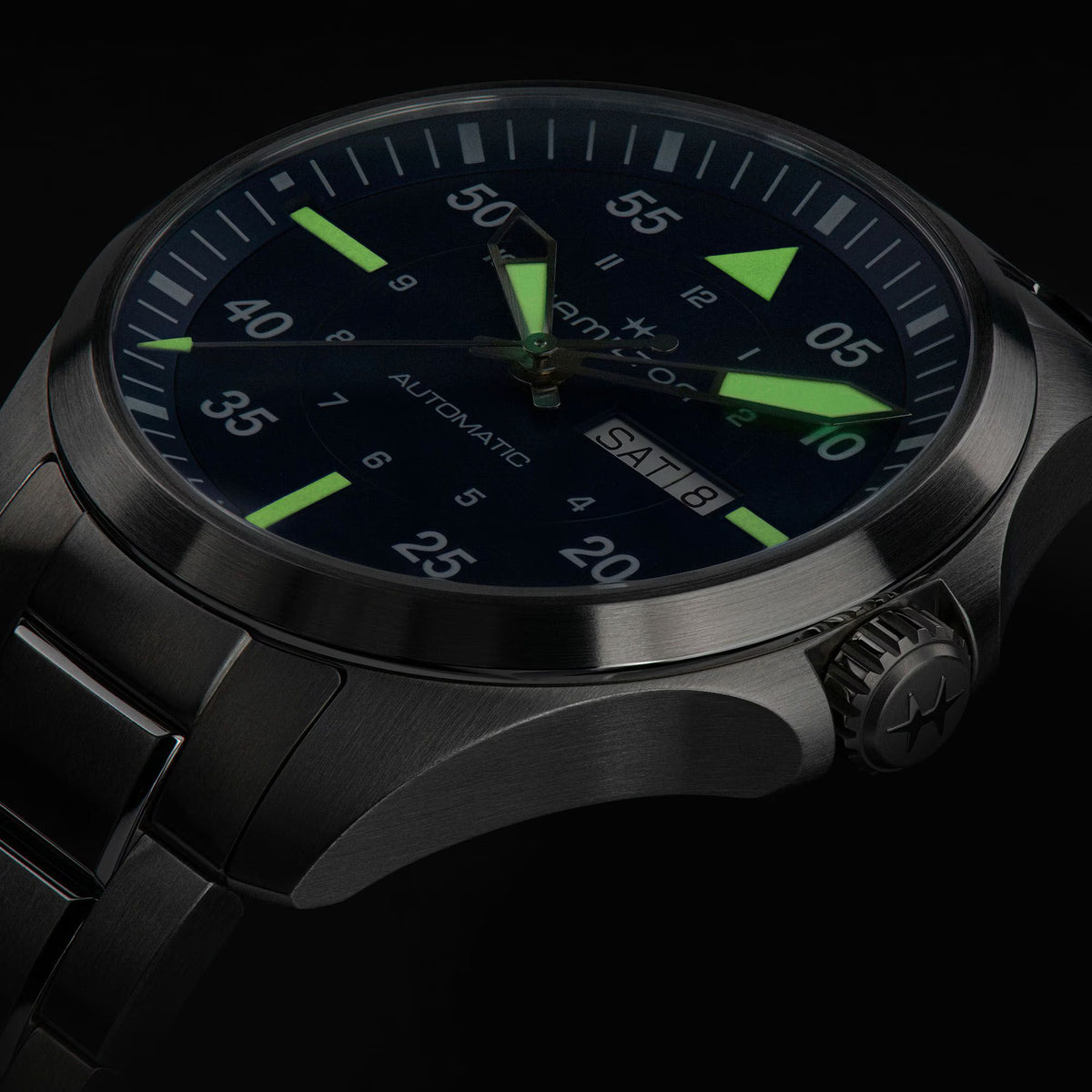 Close-up of a dark grey Hamilton automatic watch highlighting its glowing green luminous hands and hour markers in low light