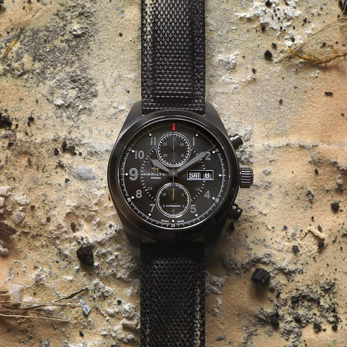Hamilton Khaki Field H71626735 top-down studio view laid flat on a textured, sandy surface to showcase its tactical aesthetic