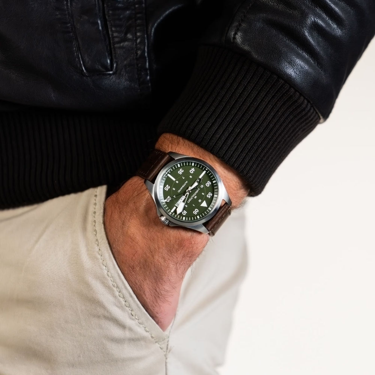 Hamilton Khaki Aviation H64635560 automatic watch wrist shot styled with a leather jacket, showing a green dial and brown leather strap
