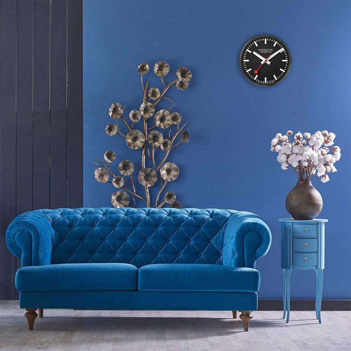 Mondaine black face wall clock mounted on a blue interior wall above a blue tufted sofa