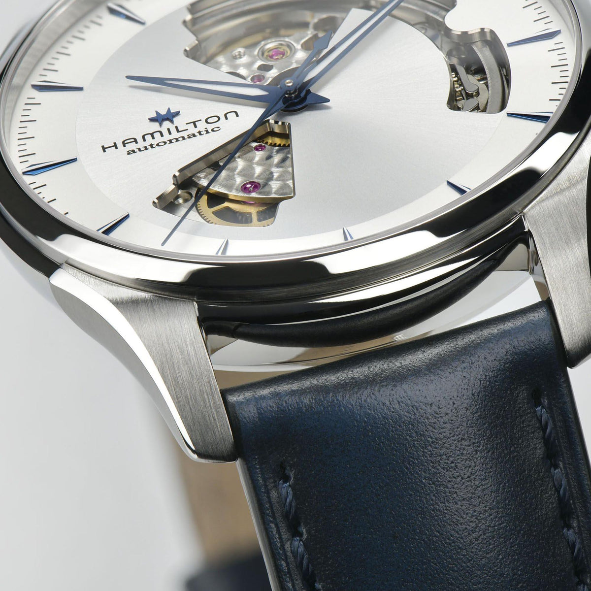 Open-heart watch close-up showing white dial, blue accents, and silver case with leather strap