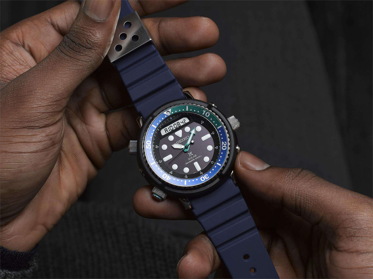 Men's dive watch held in hand, black dial, blue bezel, green markers, blue silicone strap.