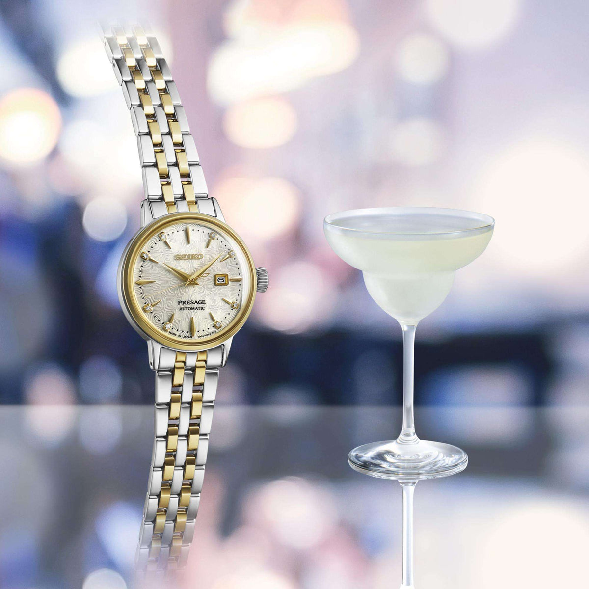 The two-tone cocktail watch presented standing vertically next to a filled margarita cocktail glass on a reflective bar counter with a blurred background.