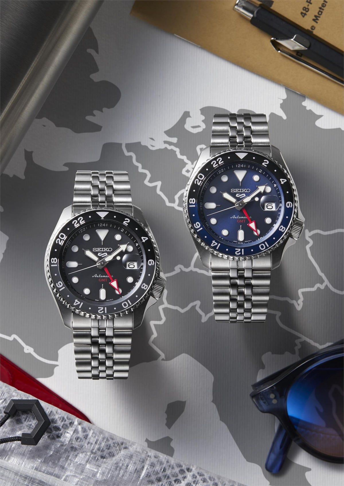 Two men's GMT watches with steel bracelets on a world map, black and blue dials shown