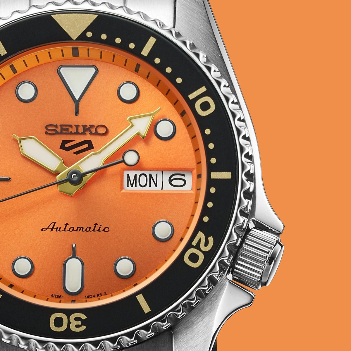 Orange dial detail with Seiko logo, gold hands, and MON 6 date window on steel case