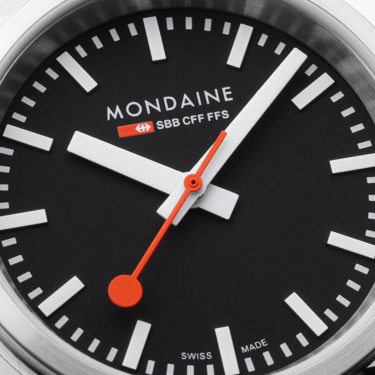 Close-up of black dial with MONDAINE logo, orange hands, and white hour markers