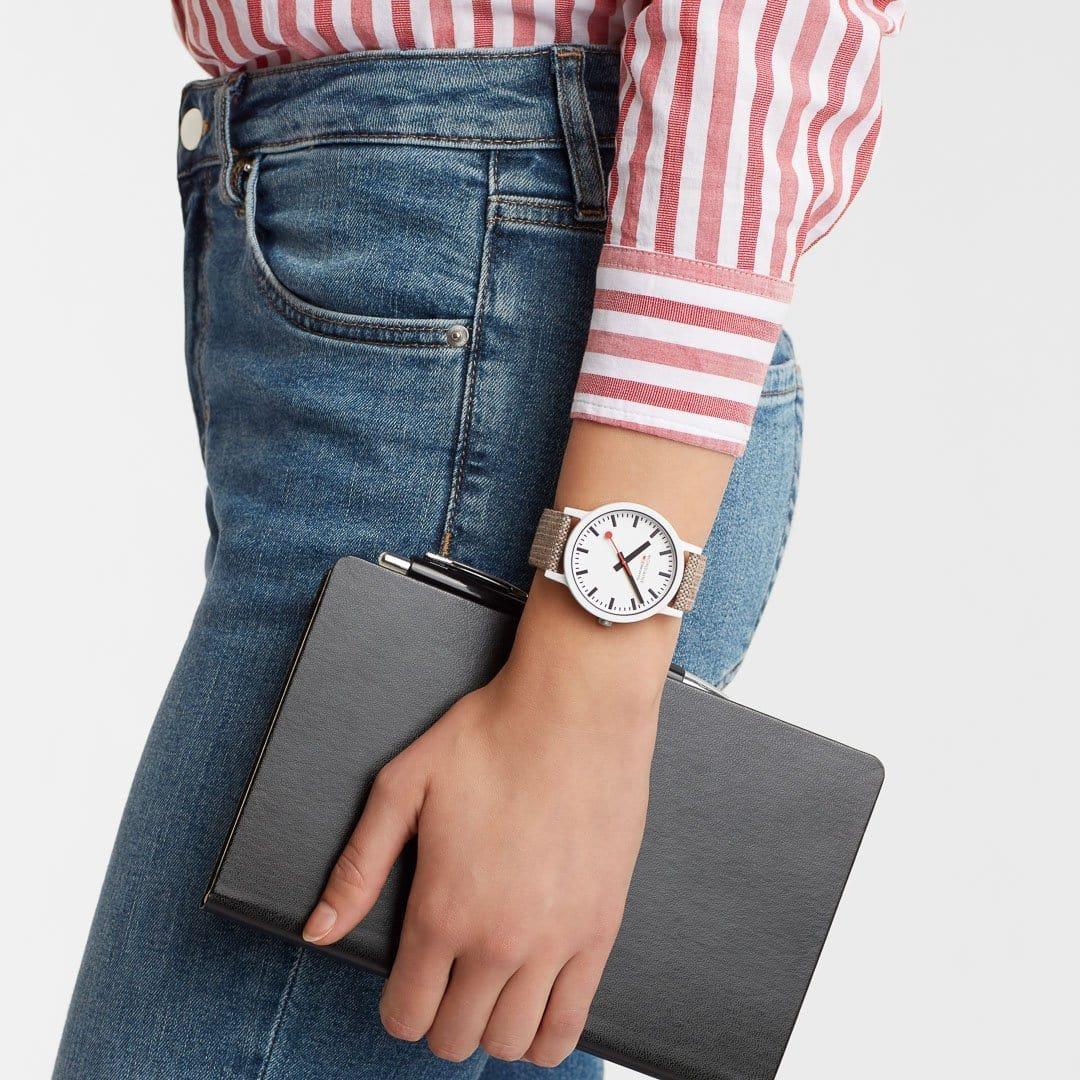Quartz wristwatch on wrist: white dial with red hands and brown textile strap.