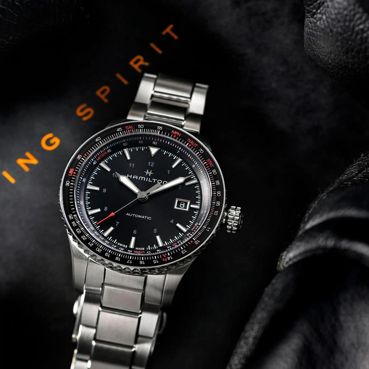 Hamilton Khaki Aviation watch styled on a black textured surface next to a leather jacket with orange text in the background