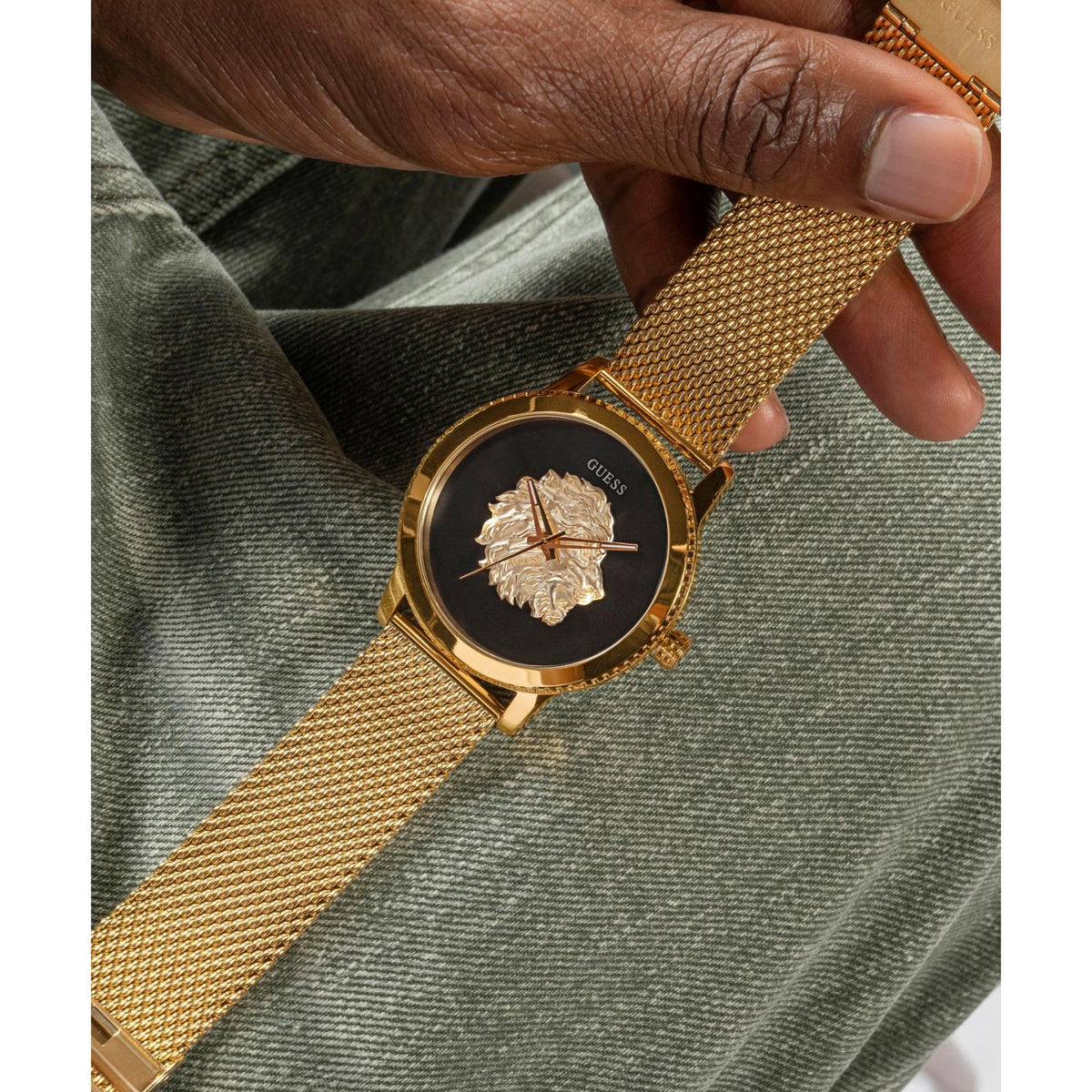 Gold-tone analog men's watch worn on wrist, black dial with lion medallion, mesh band