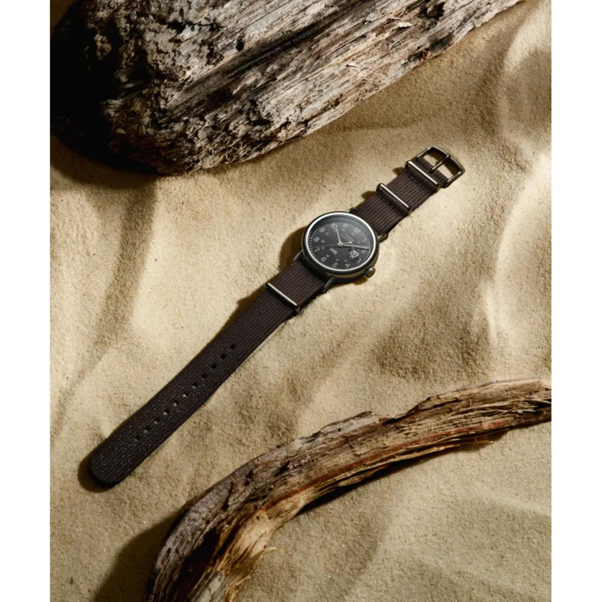 Black dial wrist watch with black fabric strap, lying on tan textured surface