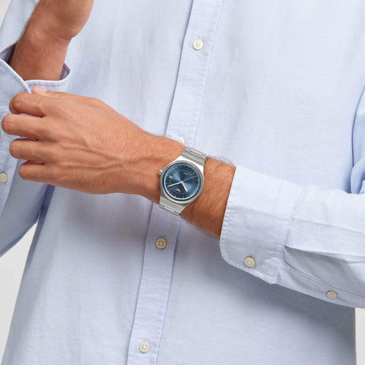 Stainless steel wristwatch on wrist over a light shirt; blue dial with date window.