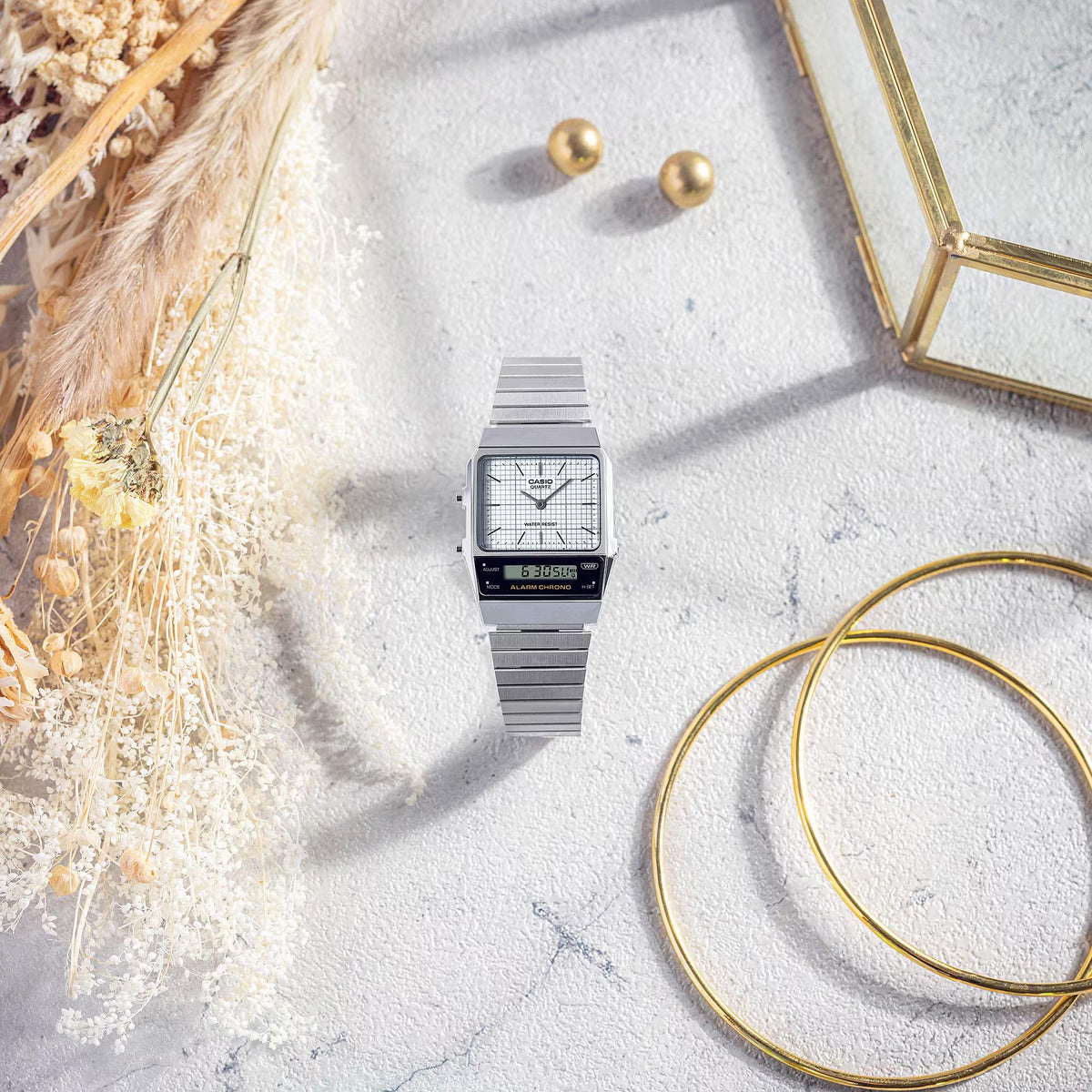 Casio Vintage Edgy AQ-800E-7AEF watch arranged on a textured light surface beside decorative dried flowers and gold accessories