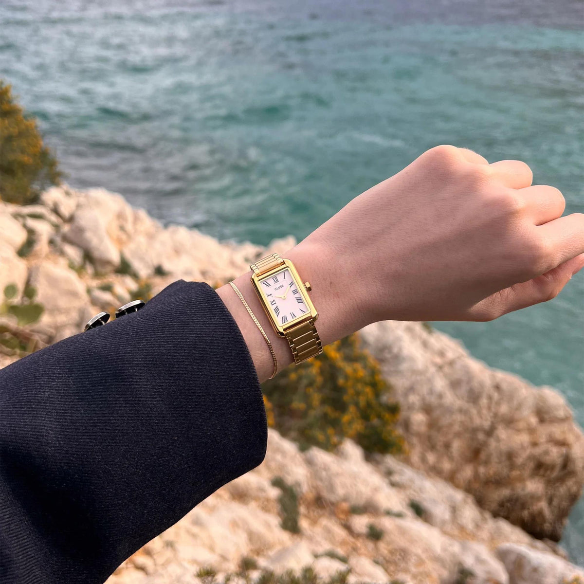Gold-tone rectangular watch on wrist with white dial and roman numerals; metal link bracelet