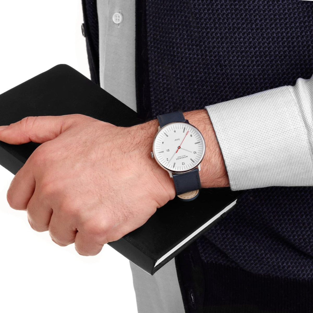 Mondaine Doppio Solar watch MLE.41910.LDV with a silver case and blue strap, worn by a person in a blue patterned knit sweater holding a black book