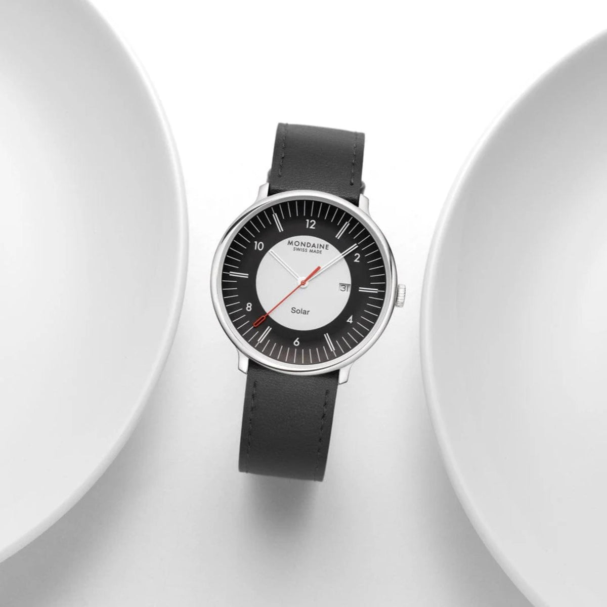 Top-down view of the Mondaine Doppio MLE.41920.LBV Solar watch with its black leather strap, artfully framed between the curved white edges of two ceramic plates