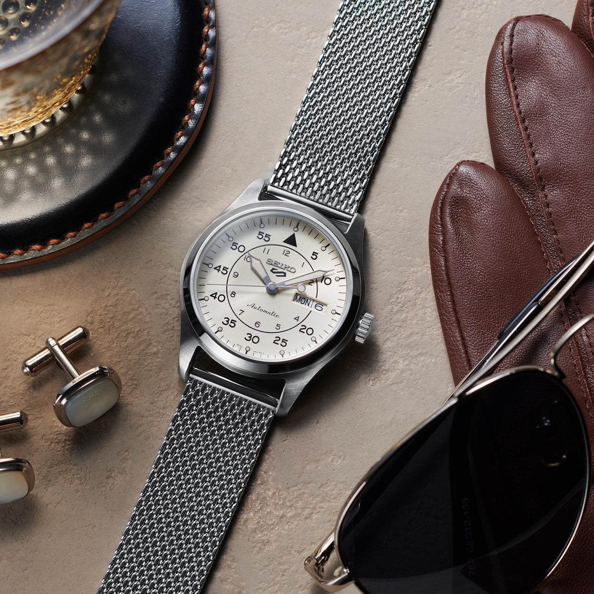 Watch with silver mesh strap on a textured surface with sunglasses, gloves, and cufflinks.
