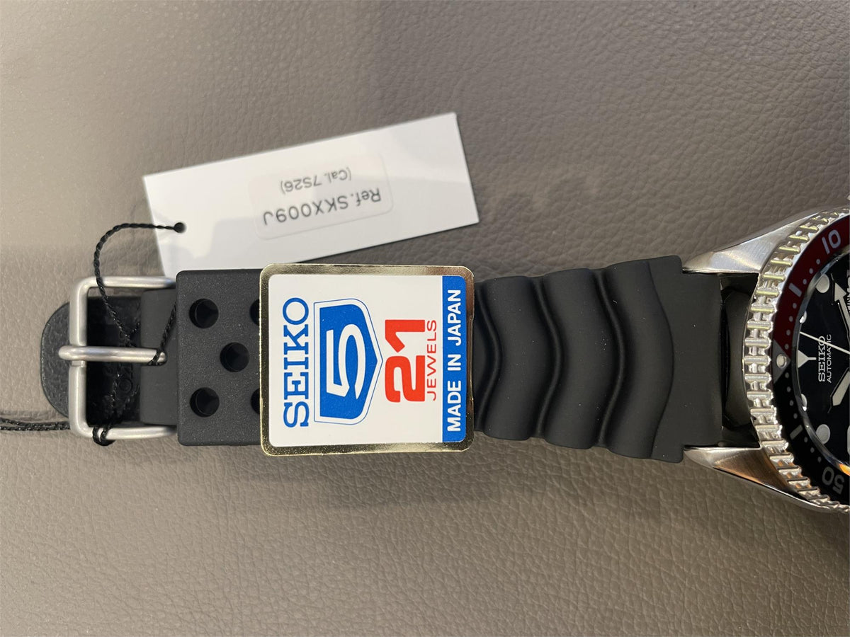 Seiko Diver’s Pepsi black rubber strap, showing the steel buckle, attached paper tag reading "Ref. SKX009J", and a blue-and-red sticker badge.