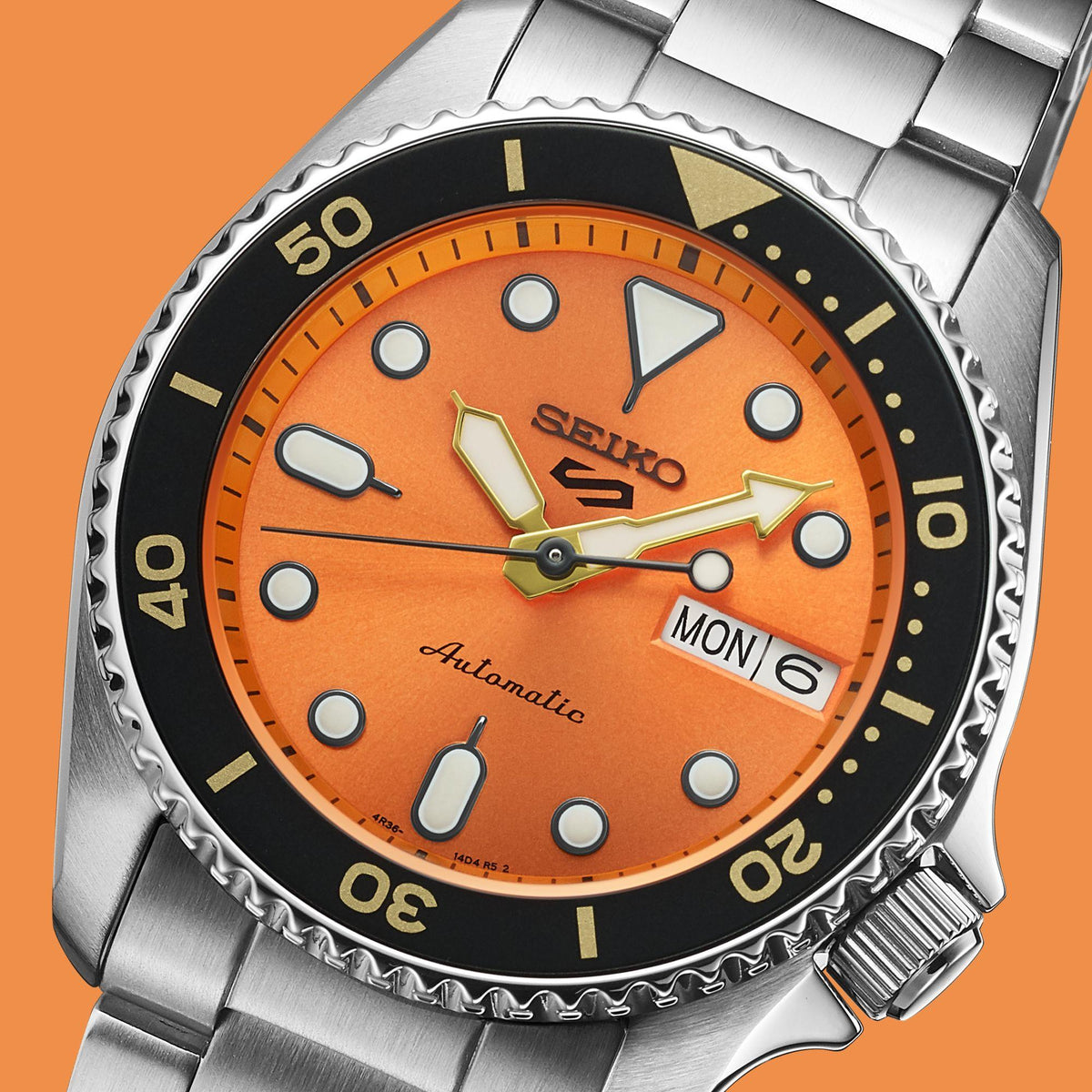 Orange dial close-up with black bezel, gold markers, and MON 6 date window