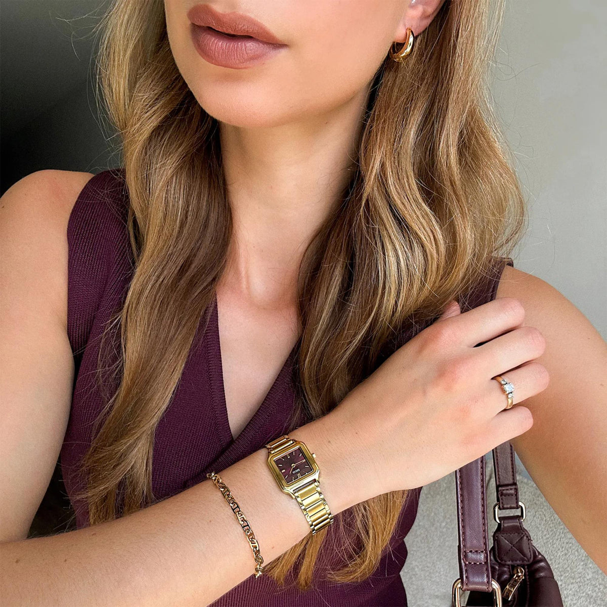 Gold metal watch with burgundy dial and roman numerals on wrist over maroon top