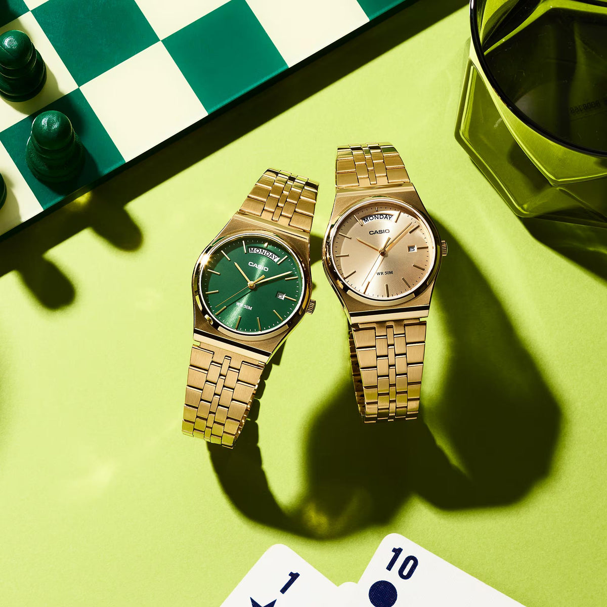Casio Standard MTP-B146G-3AVEF flat lay collection view showing the green dial model next to a gold-dial variant on a lime green surface with a chessboard and playing cards