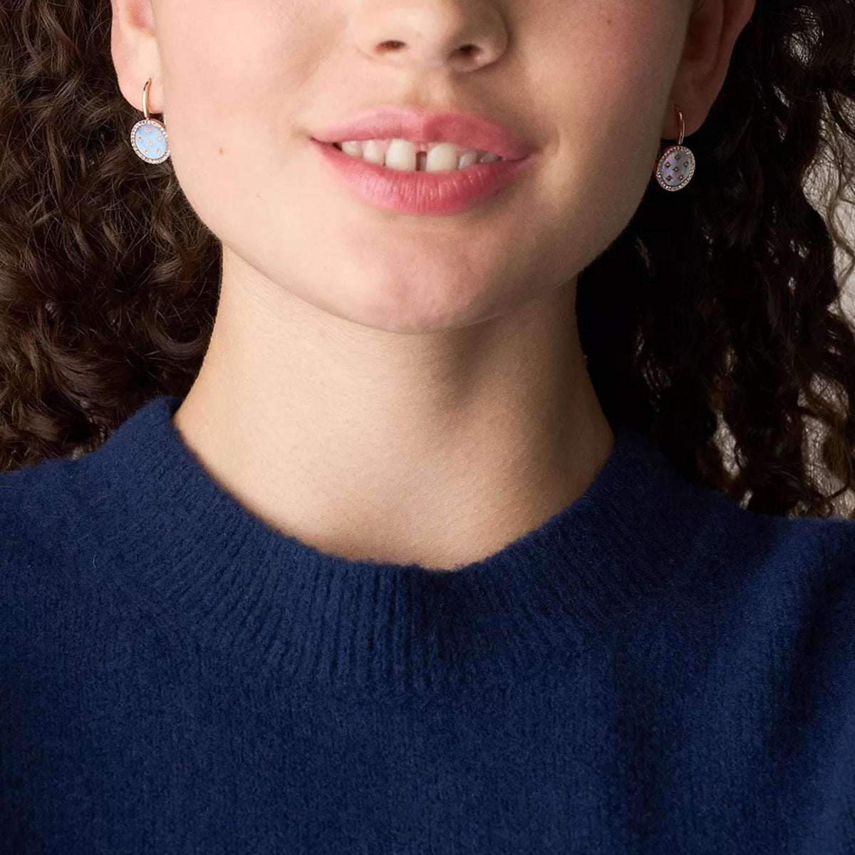 Close-up of model wearing oval blue iridescent drop earrings with crystal halo and rose gold hooks.