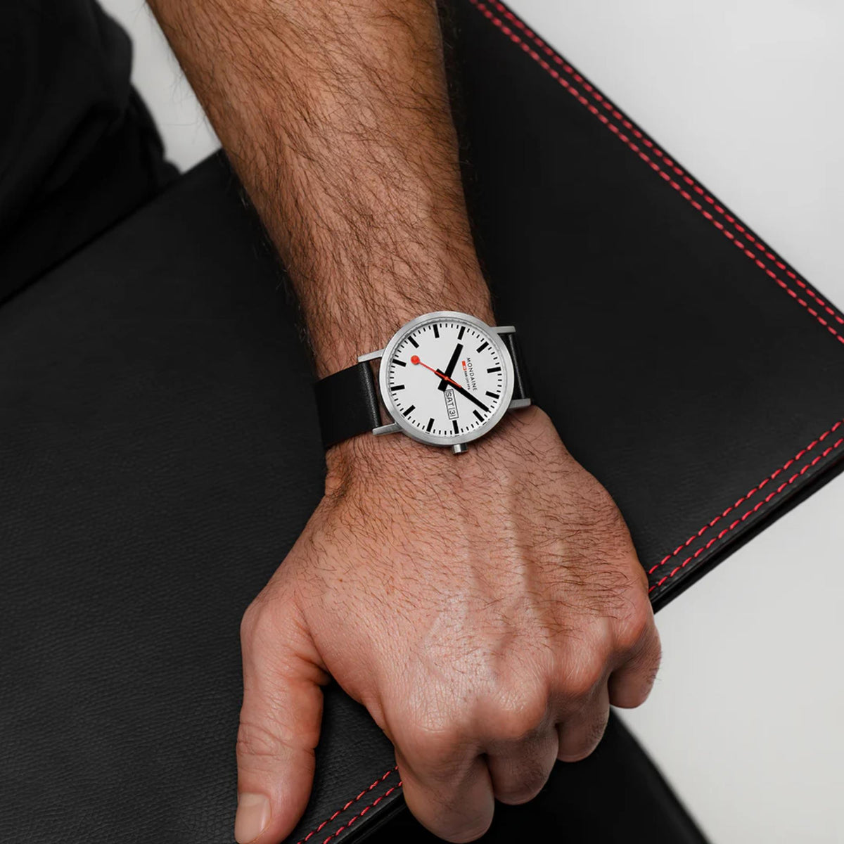Mondaine Classic A667.30360.11SBBV 40mm quartz wrist watch worn on a mans arm, resting on a black leather folder
