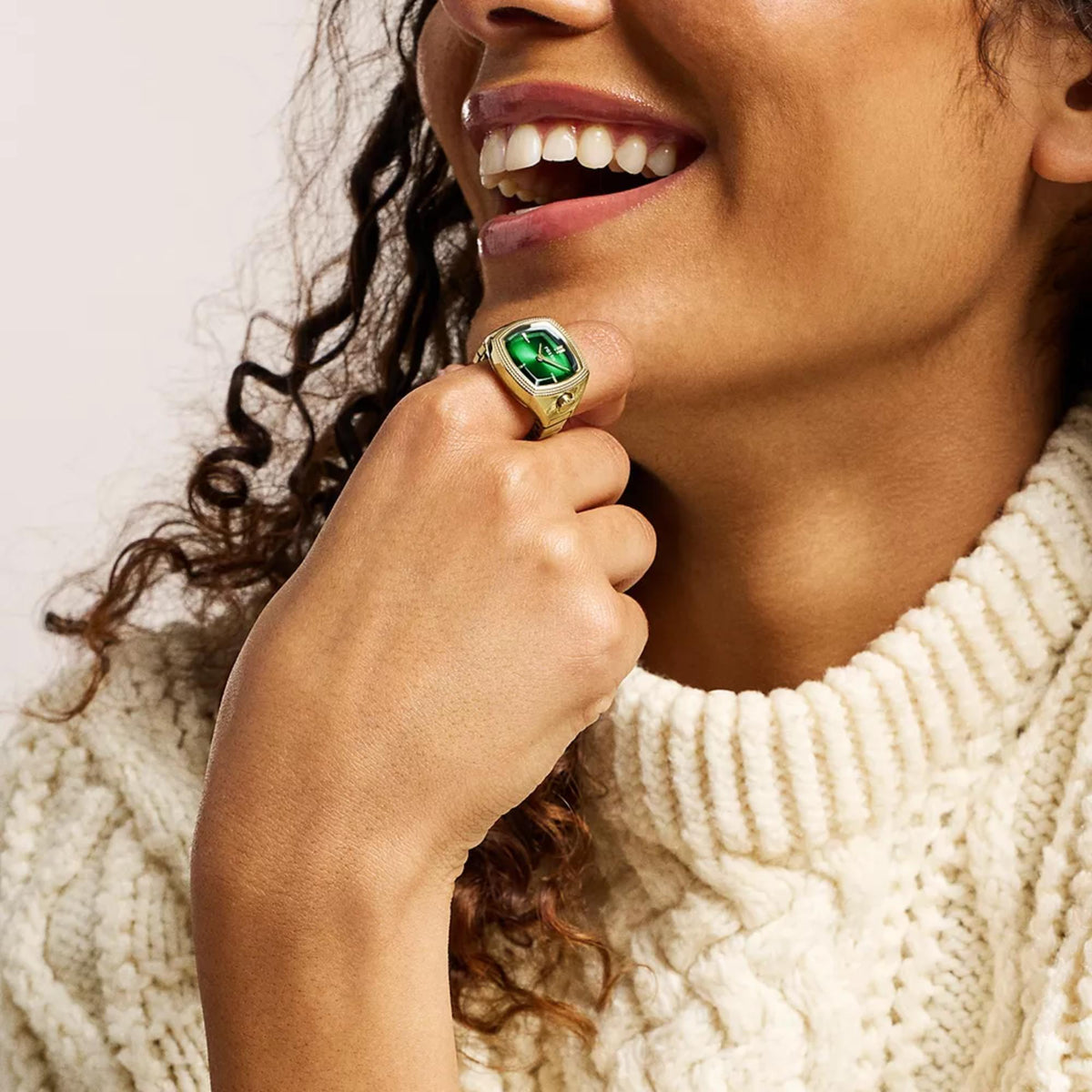 Gold ring watch with green face on finger, shown with knitted sweater sleeve