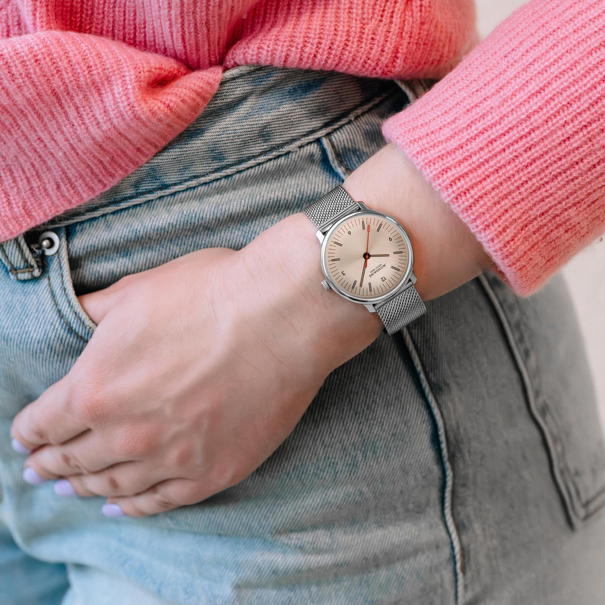 Mondaine watch worn on a wrist, paired with jeans and a pink sweater, highlighting its slim case and elegant profile in daily wear