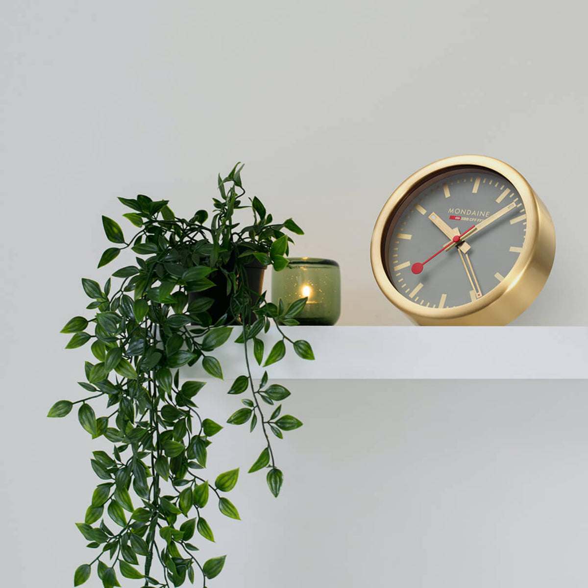 Mondaine brushed brass table clock with gray dial styled on a white floating wall shelf