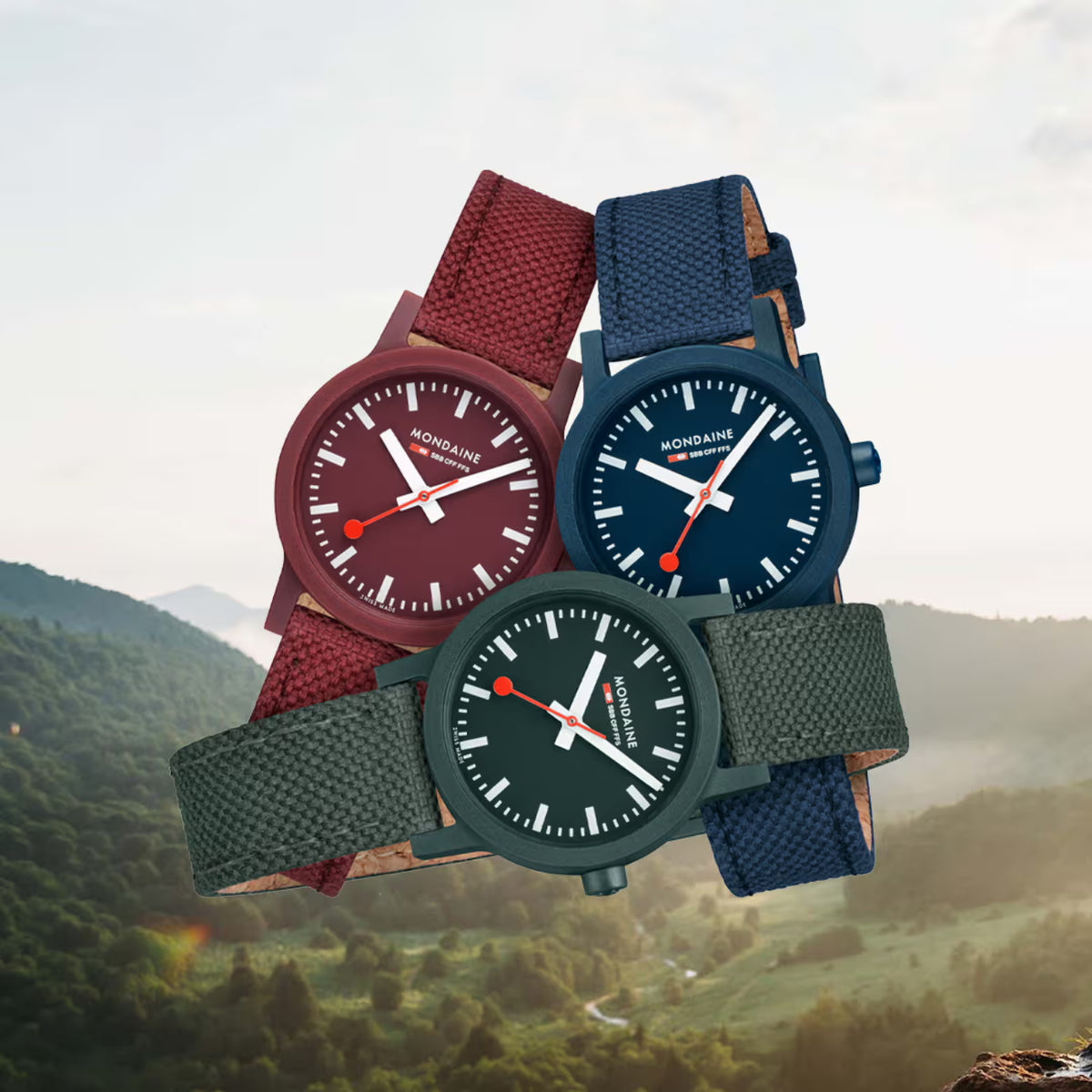 Three Mondaine eco-conscious wristwatches in burgundy, navy blue, and forest green with textile straps