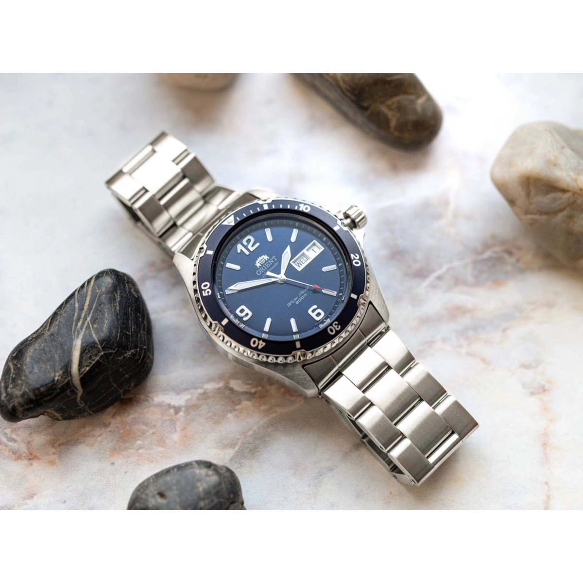 Men's steel automatic watch with blue dial and clasp, laid on rocks with bracelet extended