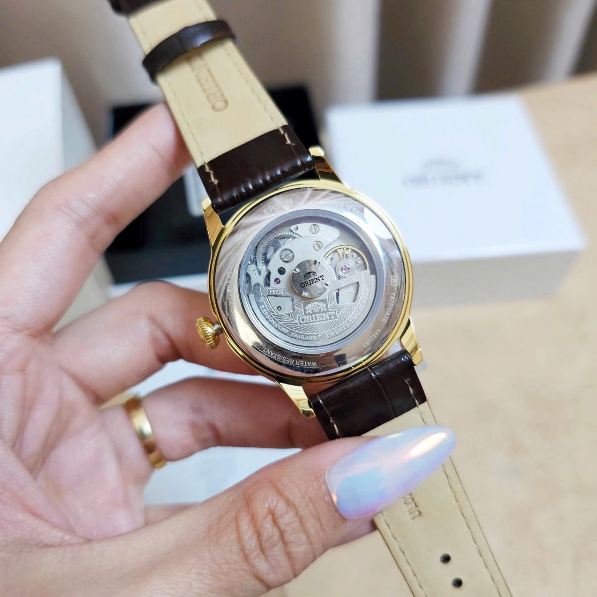 Hand holding Orient Bambino Version 7 RA-AC0M01S30B, a gold-tone wristwatch showing its exhibition caseback and a visible brand name