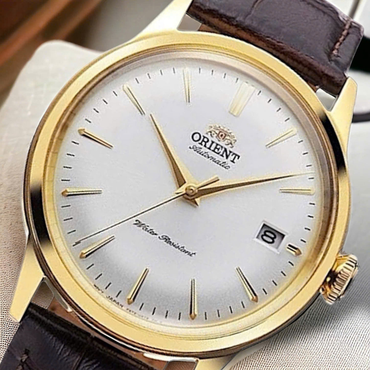 Orient Bambino RA-AC0M01S30B gold-framed wristwatch with white dial and brown leather strap on a light fabric background