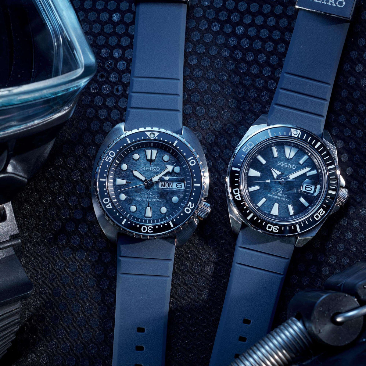 Two men’s Seiko diver watches with blue silicone straps on dark patterned surface