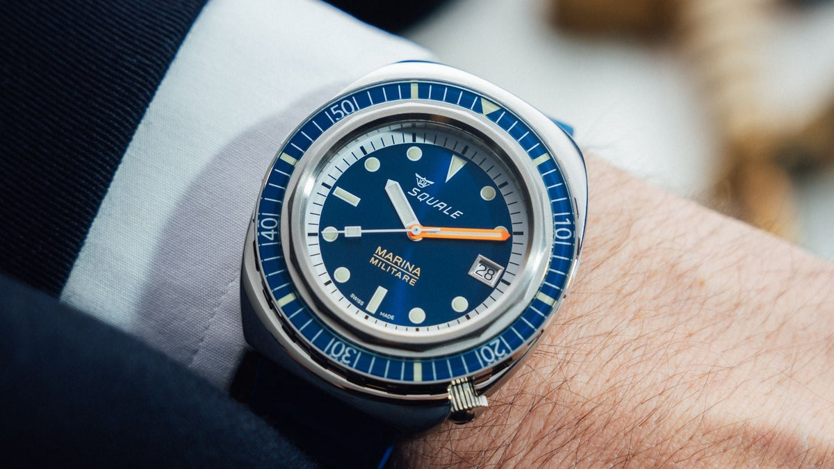 Close-up of the blue Squale dive watch worn on a wrist, showcasing its orange hand and unique 4 o'clock crown next to a suit cuff